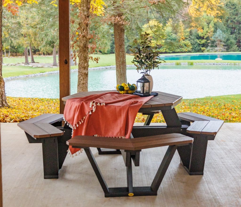 Brown and black poly lumber Octagon Picnic Table decorate with a red table linen, a black lantern, a bowl of lemons and limes, and a plant in a white pot sits in front of a small pond.