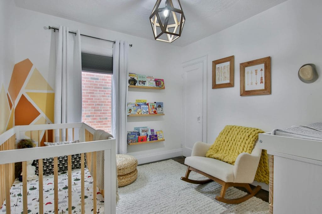 Nursery with white walls, an orange and yellow wall painting, a white wood crib, a white rocking chair with a yellow pillow, and three shelves full of baby books.