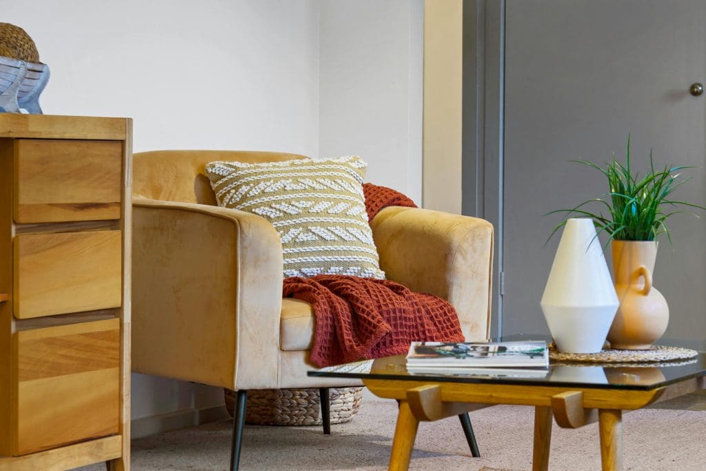 Yellow velvet armchair with dark orange blanket and white patterned throw pillow with a coffee table and shelves next to it.