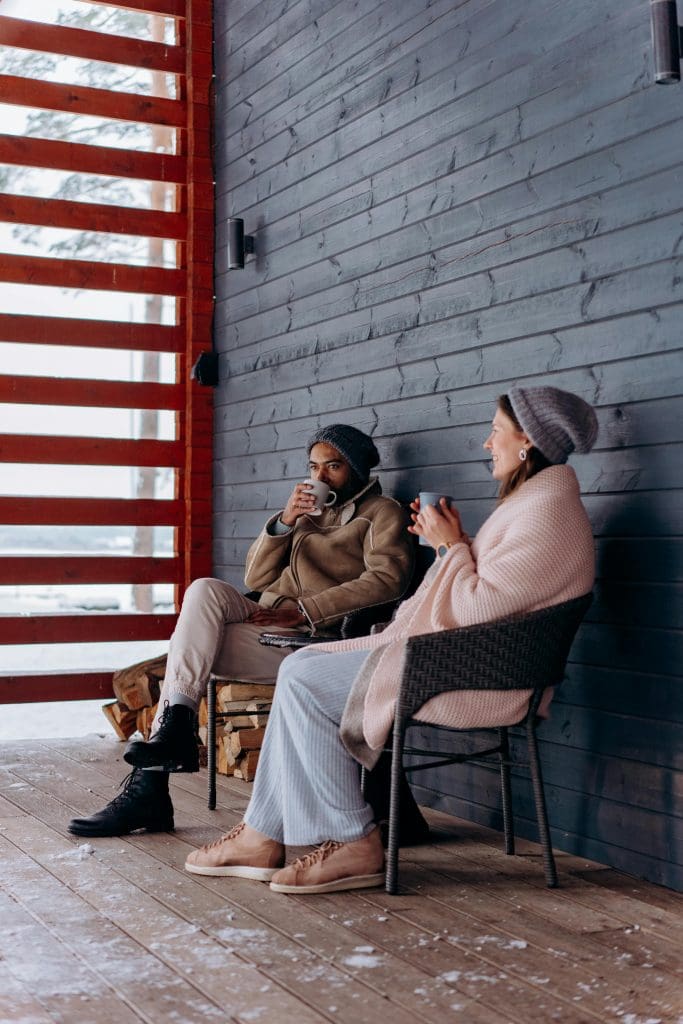 A couple sits on their porch bundled up during the winter time drinking from mugs.