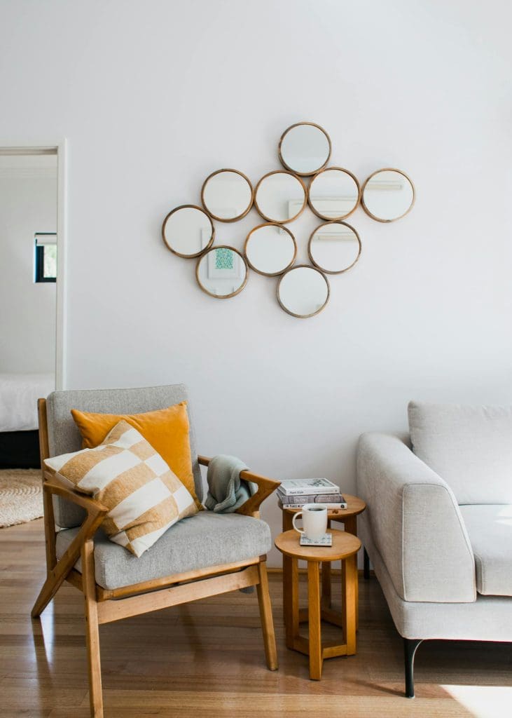 Gray and hardwood chair with yellow pillows, a white couch, 2 small round end tables, and a collage of circle mirrors.