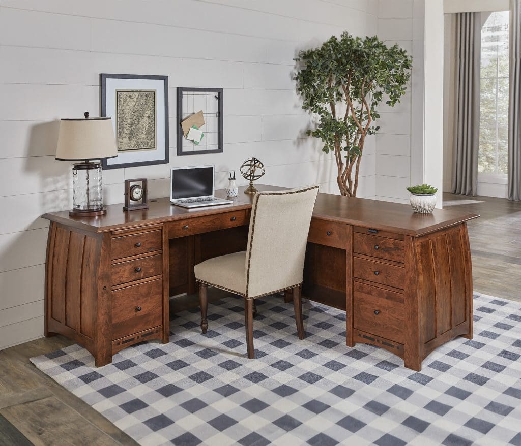 A home office with a brown hardwood desk with curved sides, a white upholstered chair, a plaid rug, industrial decor, and 2 plants.