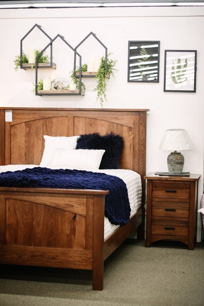 Brown wood bed with white and blue bedding and matching nightstand in a room with tan carpet, white walls, and plant decor.