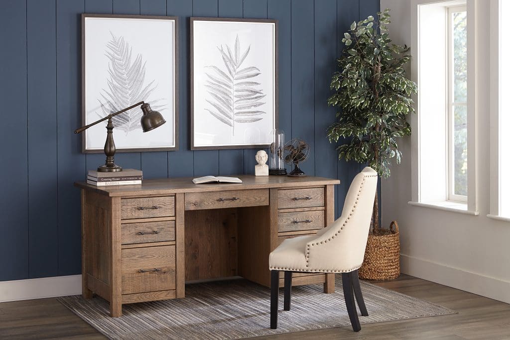 Wood desk with 7 drawers in a room with a dark blue accent wall, fake tree plant in a corner, white walls, wood plank flooring, a cream office chair, 2 large leaf prints, and a brown and white rug.