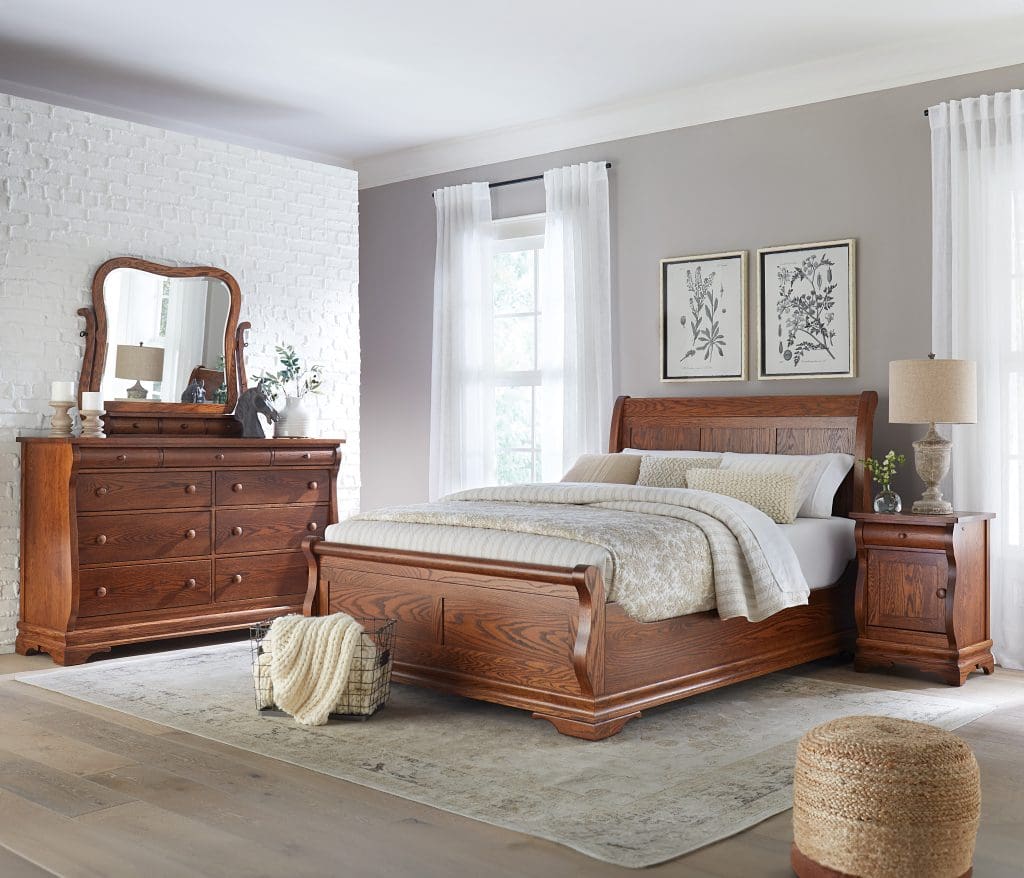 Bedroom with brown hardwood sleigh bed frame with white bedding, a brown hardwood long 6-drawer dresser with a curved mirror, light gray walls, light hardwood flooring with a large white rug, and windows with white sheer curtains.