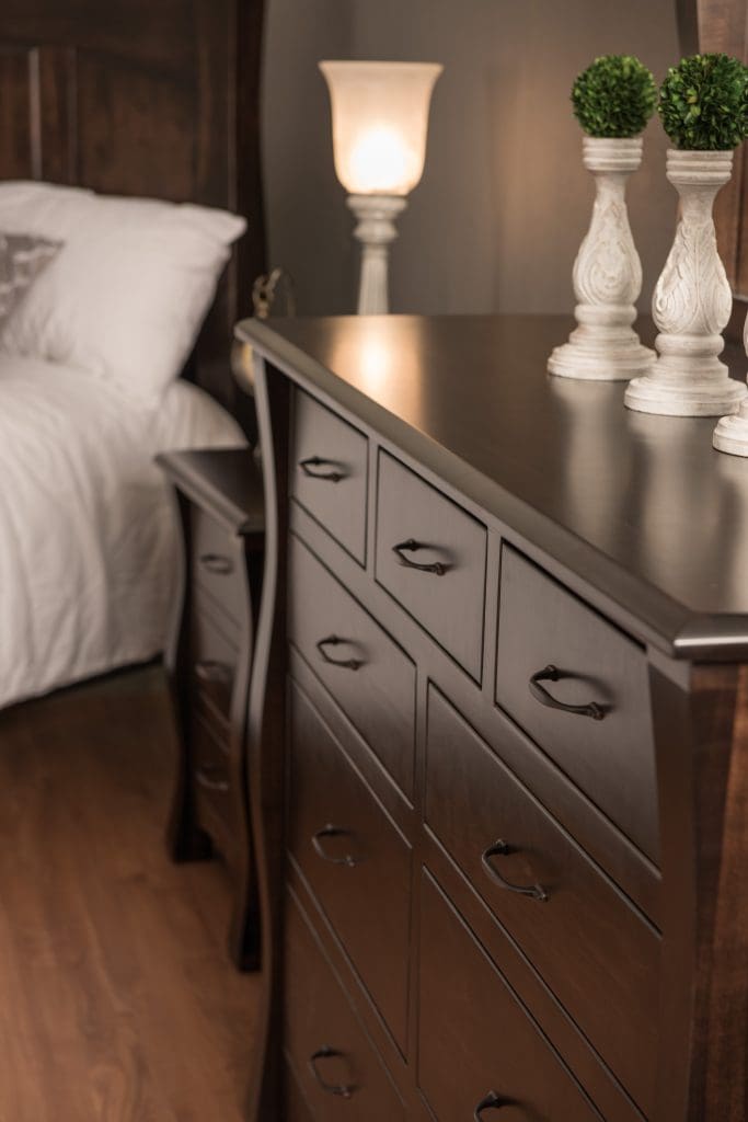 Close up of a dark brown hardwood dresser with a matching nightstand and bed in the background.
