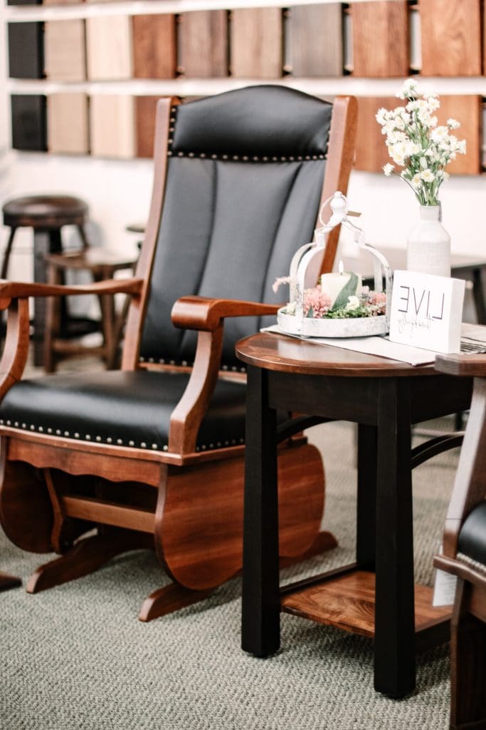 Close up of a wooden glider rocker with a black leather upholstery and sitting next to a two toned black and wood end table with a collection of home decor on it.