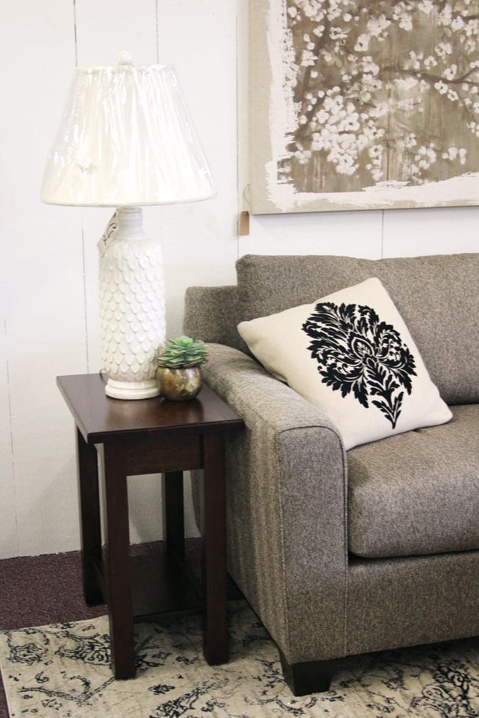 Close up of a light gray couch with a white and black pillow and sitting next to a dark brown end table with a large white lamp.