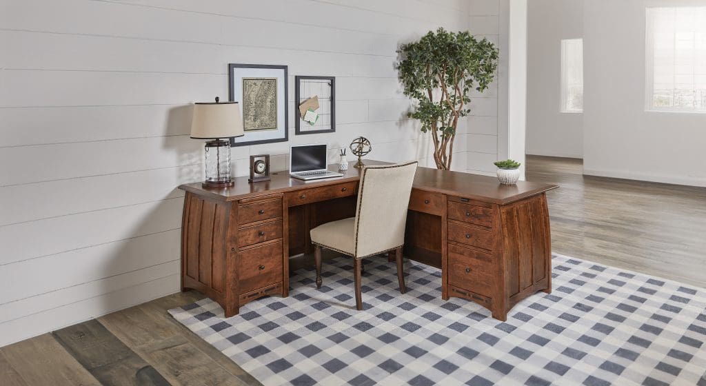 A home office with a brown hardwood desk with curved sides, a white upholstered chair, a plaid rug, industrial decor, and 2 plants.