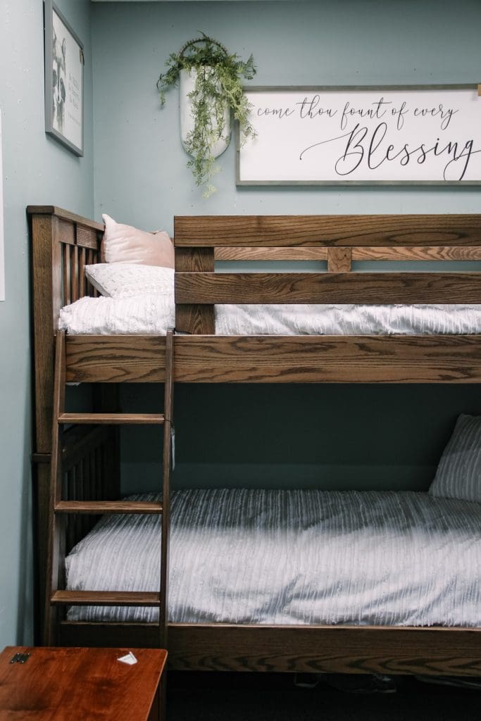 A light blue room with a oak wooden bunkbed with a White bed spread and white pillows, and a gray framed word artwork on the wall. 