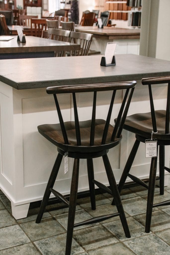 A kitchen scene with laminate floors and hardwood furniture in the background. Has a white island with a grey concrete top, with quality swivel counter stools in a mid century style