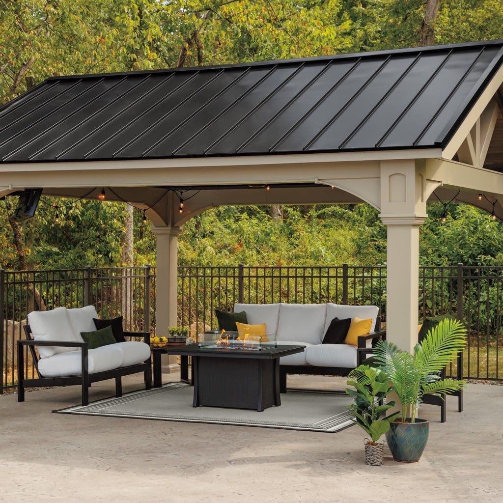 Black rectangle fire table in coffee table height with coordinating black and white poly furniture outdoor sofas around it on a deck with a pergola over it. 