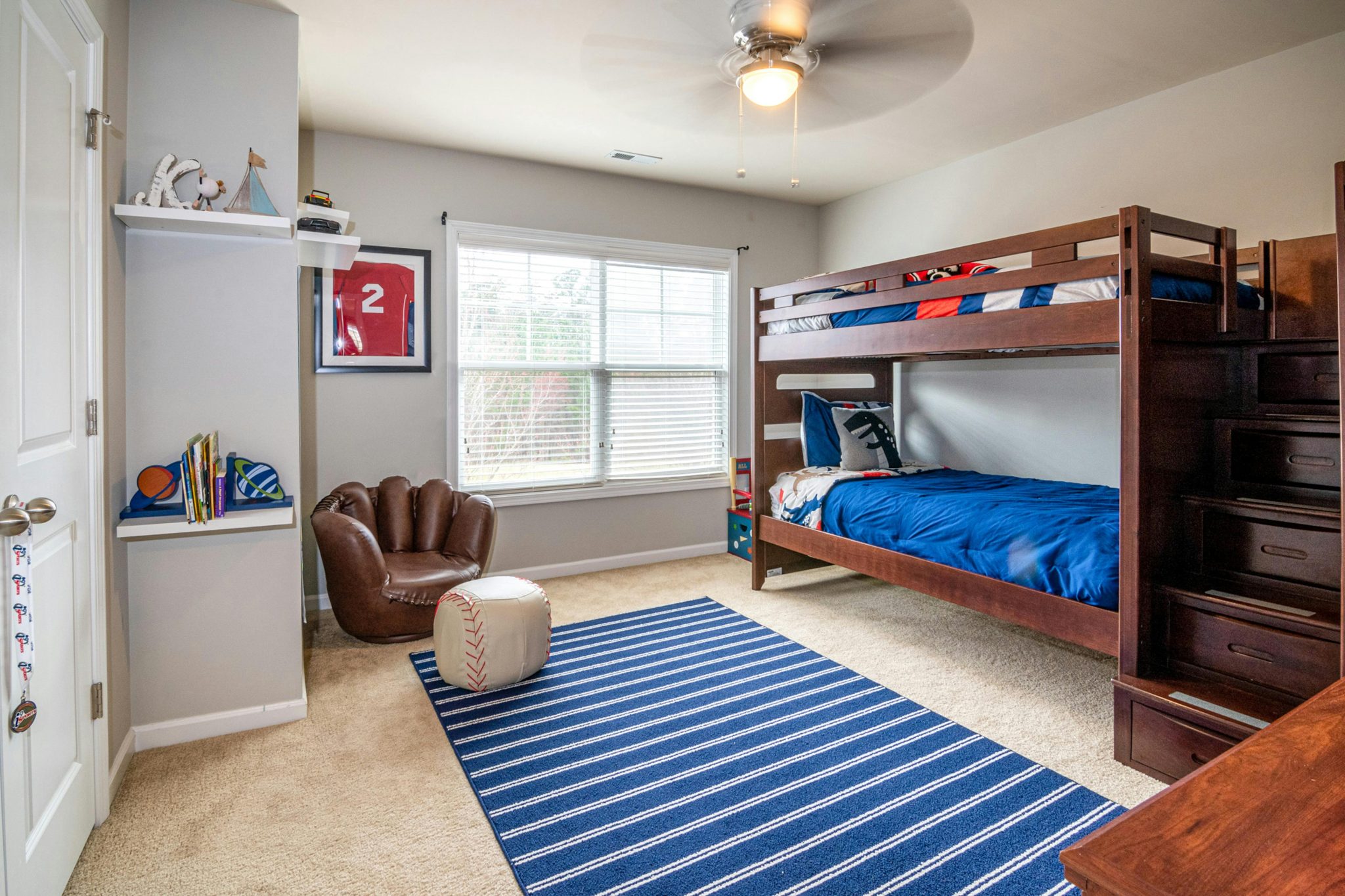 Sports-themed kid's room with white walls, a large blue and white rug, and brown wood bunk beds with blue bedding and stairs.