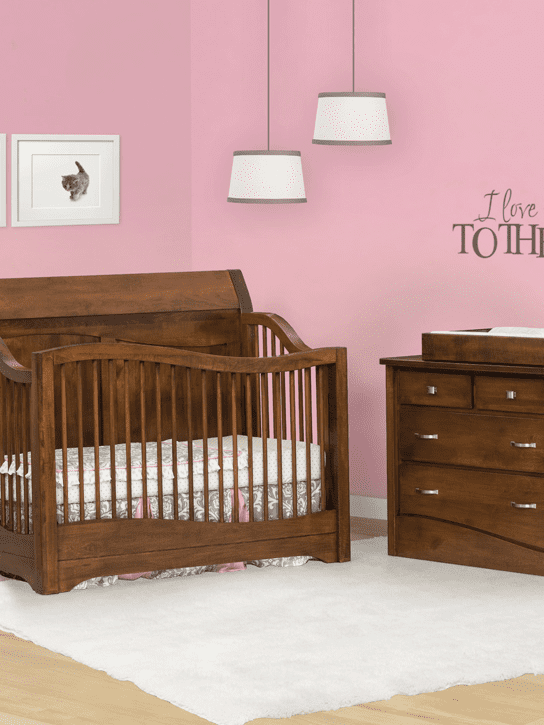 A pink nursery with light wood floors and a rectangle white rug. with the wave panel crib in dark hardwood on the back, sides and front. And a dark hardwood changing table.